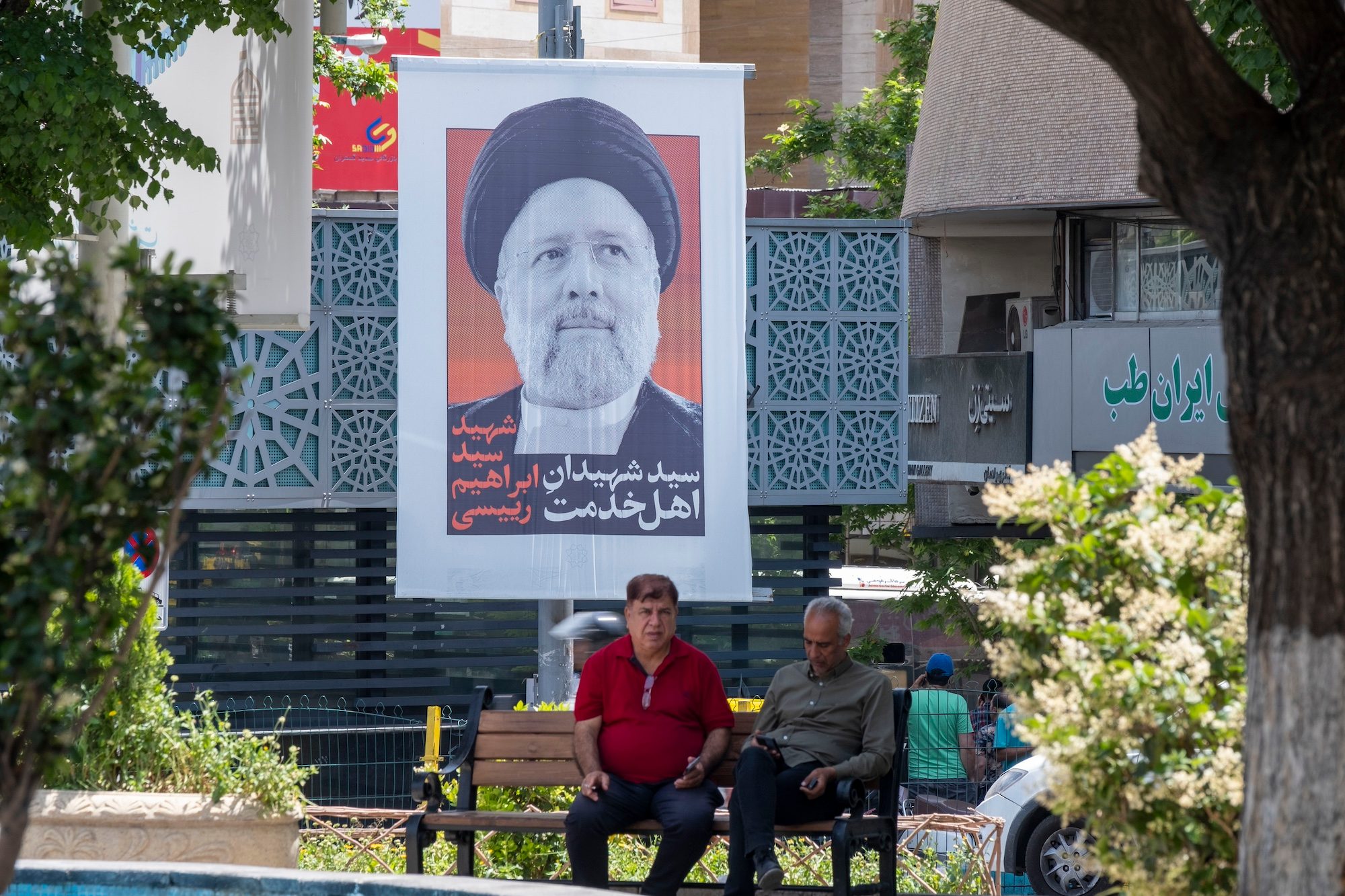 Two men sit beneath a tribute to President Ebrahim Raisi in Tehran; Iran has said it will increase exports but analysts speculate that the announcement my be politically motivated