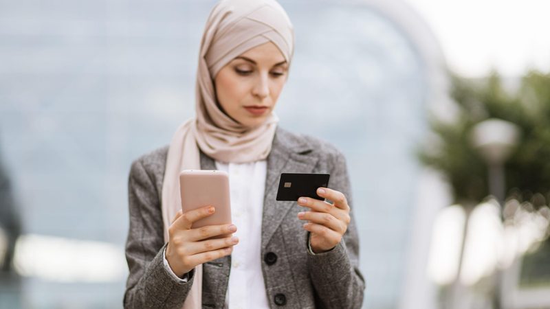 Pretty Muslim lady in hijab using smartphone and credit bank card for online shopping. Focused businesslady in headscarf standing on street and making purchase.