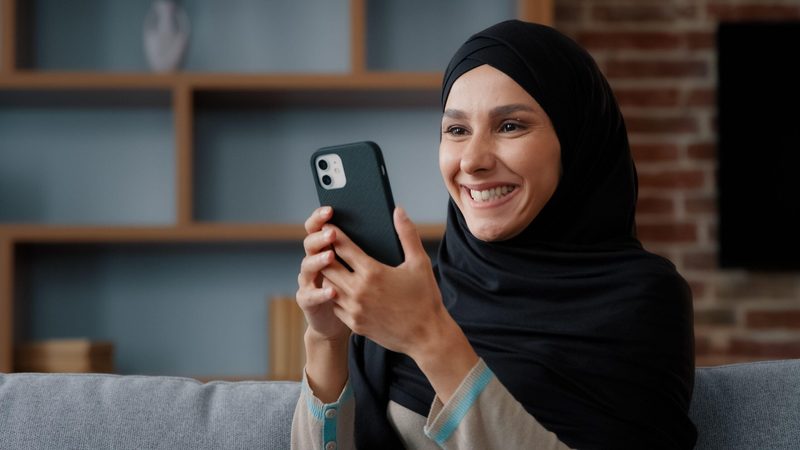 Smiling Arab Muslim Islamic 30s woman user in headscarf black hijab using phone at home sitting on couch middle eastern girl lady holding mobile