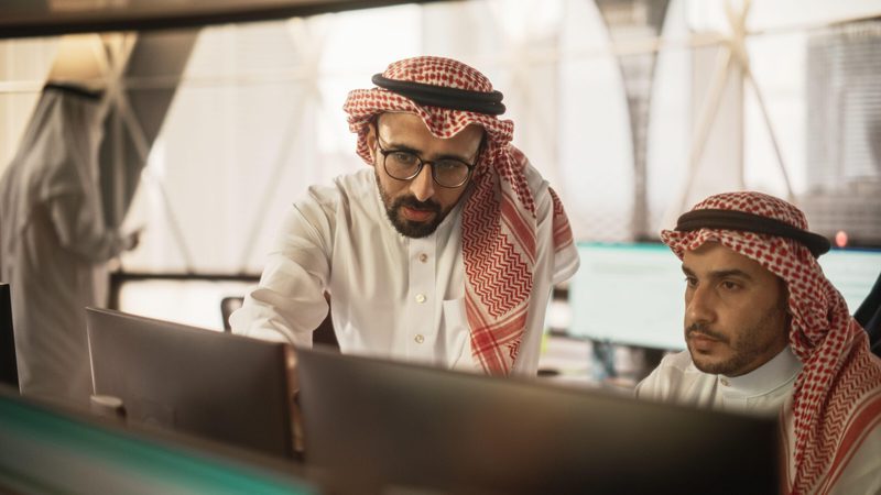 Arab Graphic Designer Working on Computer, Discussing Project with Team Leader. Middle Eastern Colleagues Working in Modern Office, Wearing Traditional White Thobes and Kaffiyeh