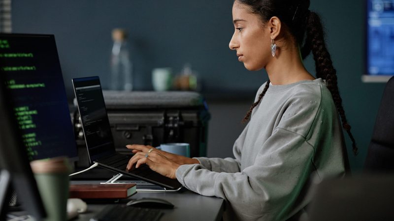 Side view of professional female IT developer from Middle East coding at ease on laptop while sitting at workstation with several computers in office