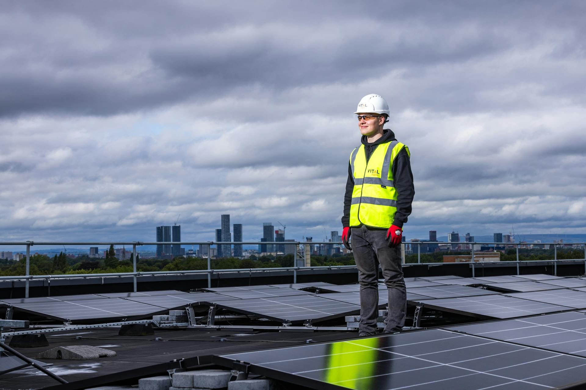 A man in a high-vis vest and white hard hat standing among solar panels on a roof with a cloudy sky and city skyline in the background. Renewables such as solar power are creating an energy addition, not a transition, says historian Daniel Yergin